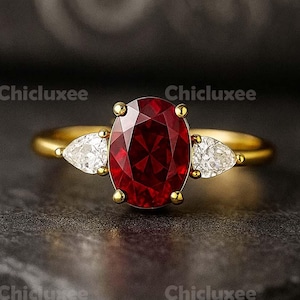 Könnte beinhalten: Ein goldener Ring mit einem großen, oval geschliffenen, tiefroten Edelstein, flankiert von zwei birnenförmigen, weißen Edelsteinen. Der Ring hat ein klassisches Drei-Stein-Design, vor dunklem Hintergrund.