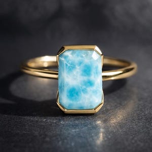 Op de afbeelding: Een gouden ring met een rechthoekige, lichtblauwe edelsteen met witte adering. De edelsteen is gezet in een gouden bezel en de ringband is een dun, gepolijst goud. De ring wordt tegen een donkere achtergrond getoond.