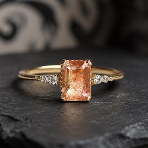 Peut inclure: Une bague en or ornée d'une pierre précieuse rectangulaire de couleur pêche. De petites pierres précieuses claires sont serties de chaque côté de la pierre principale. La bague est présentée sur une surface sombre et texturée, avec un fond à motifs.