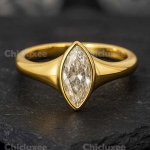 May include: A gold ring with a marquise-cut diamond center stone. The ring has a polished gold band and a bezel setting that holds the diamond. The ring is displayed on a dark, textured surface. The word "Chicluxee" is visible in the image.