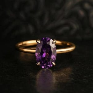 May include: A gold ring featuring a central oval-cut purple gemstone held by four prongs. The band is a smooth, polished gold metal. The gemstone displays deep violet hues and multifaceted surfaces that catch the light, centred on the band.