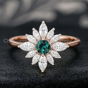 Pode incluir: Anel em ouro rosa com design floral. A pedra central é uma gema verde escura redonda, rodeada por gemas brancas em forma de marquesa. A banda do anel é fina e em ouro rosa polido. O anel está sobre um fundo escuro.