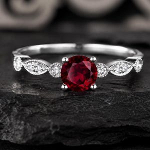 May include: A silver-toned ring featuring a round, deep red center stone held by four prongs. The band is accented with small, clear, round and marquise-shaped stones set in a delicate, milgrain-edged metal frame along the sides of the center stone.
