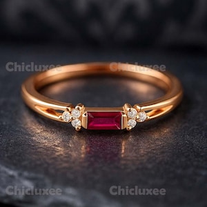 May include: A gold ring featuring a rectangular, deep red gemstone, accented with small clusters of white stones. The ring's slender band and the dark, textured backdrop create a striking contrast.