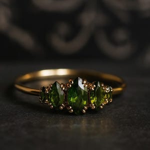 Op de afbeelding: Een gouden ring met vijf marquise-geslepen groene edelstenen. De ring heeft een slanke band en de edelstenen zijn gezet in een decoratieve gouden zetting. De ring wordt gepresenteerd op een donkere ondergrond.