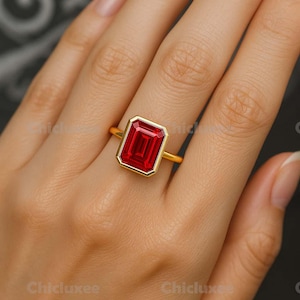 Könnte beinhalten: Ein goldener Ring mit einem großen, rechteckigen, roten Edelstein. Der Edelstein ist in eine goldene Lünette gefasst. Der Ring befindet sich an einem Finger. Der Ring ist ein Schmuckstück.