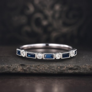 May include: A silver ring featuring alternating rectangular blue and round clear gemstones. The ring is displayed on a dark, textured surface, highlighting the detailed design and sparkle. The ring is a band style.