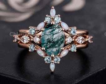 May include: A rose gold ring set featuring a large oval-cut green gemstone with white veining, surrounded by smaller opalescent and blue gemstones. The set includes a matching band with similar accent stones.