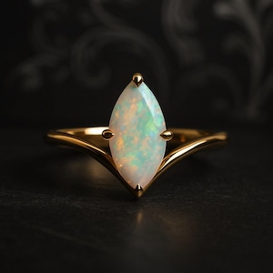 Könnte beinhalten: Ein goldener Ring mit einem Opal im Marquiseschliff. Der Opal hat eine weiße Basis mit schillernden grünen und blauen Reflexen. Der Ring hat ein zartes, V-förmiges Design. Die Fassung hat kleine goldene Akzente.