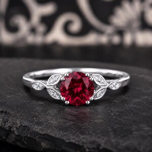 May include: A silver ring featuring a round, deep red center stone held by prongs. The band is accented on both sides of the center stone with two marquise-cut clear stones arranged in a leaf-like pattern, set into the polished silver metal band.