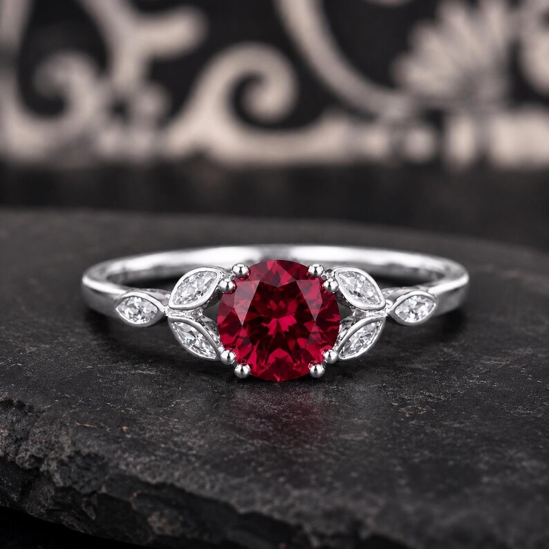 May include: A silver ring featuring a round, deep red center stone held by prongs. The band is accented on both sides of the center stone with two marquise-cut clear stones arranged in a leaf-like pattern, set into the polished silver metal band.