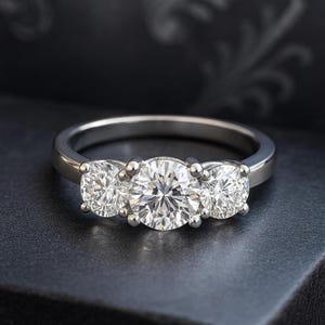 Op de afbeelding: Een driestenen verlovingsring met een zilveren band. De ring heeft drie ronde, briljant geslepen diamanten, waarbij de middelste steen iets groter is dan de zijstenen. De diamanten schitteren tegen een donkere achtergrond.