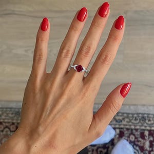 May include: A silver-toned ring featuring a round, deep red center stone flanked by two small, marquise-cut clear stones on each side, set on a slender, polished metal band that rests on a finger with bright red nail polish.
