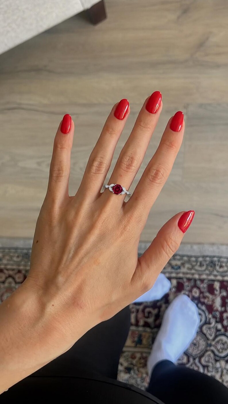 May include: A silver-toned ring featuring a round, deep red center stone flanked by two small, marquise-cut clear stones on each side, set on a slender, polished metal band that rests on a finger with bright red nail polish.