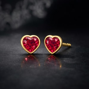 Op de afbeelding: Een paar hartvormige oorstekers. Elke oorbel heeft een levendige rode edelsteen gezet in een goudkleurige bezel. De oorbellen worden gepresenteerd op een donkere ondergrond, met een zachte, vage achtergrond.