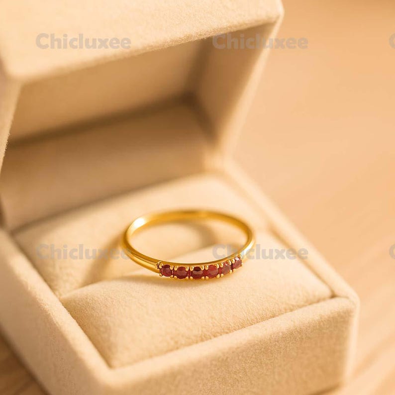 May include: A gold ring with a row of small, red gemstones set in a band. The ring is displayed inside a beige jewelry box. The ring is a delicate piece of jewelry.