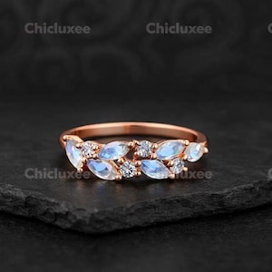 Op de afbeelding: Roségouden ring met marquise-geslepen maanstenen en ronde diamantaccenten. Het ontwerp van de ring lijkt op een bladerpatroon, met de edelstenen langs de band gezet. De ring wordt gepresenteerd op een donkere, getextureerde ondergrond.