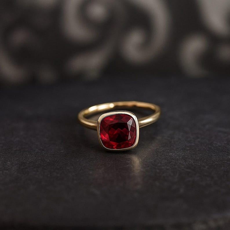 K&ouml;nnte beinhalten: Ein goldener Ring mit einem quadratischen, tiefroten Edelstein. Der Ring hat ein schlichtes, poliertes Goldband, und der Edelstein ist in einer goldenen L&uuml;nette gefasst. Der Ring wird vor einem dunklen Hintergrund gezeigt, um seine Farbe und sein Design hervorzuheben.