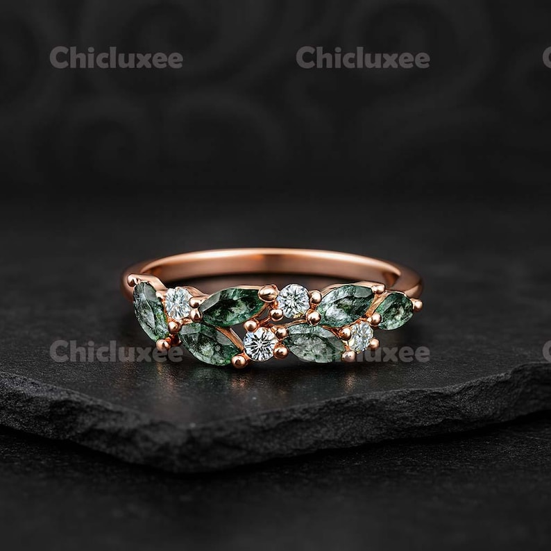 May include: Rose gold ring featuring a leaf design with green and white gemstones. The ring has a delicate band and is set on a black surface. The ring is a piece of jewelry.