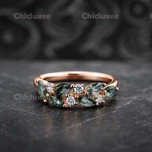 May include: Rose gold ring featuring a leaf design with green and white gemstones. The ring has a delicate band and is adorned with marquise-cut green stones and round white stones. The ring is displayed on a dark surface.