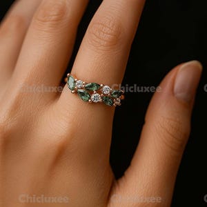 May include: A gold ring featuring a leaf-like design with green and white gemstones. The ring has a delicate, elegant appearance, with the gemstones set in a repeating pattern. The ring is shown on a person's finger.