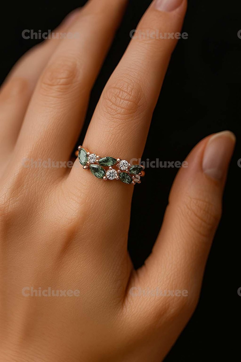 May include: A gold ring featuring a leaf-like design with green and white gemstones. The ring has a delicate, elegant appearance, with the gemstones set in a repeating pattern. The ring is shown on a person's finger.