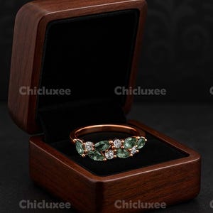 May include: A rose gold ring with green and white gemstones, displayed in an open wooden jewelry box with a black velvet interior. The box is a dark brown color. The brand name "Chicluxee" is visible.