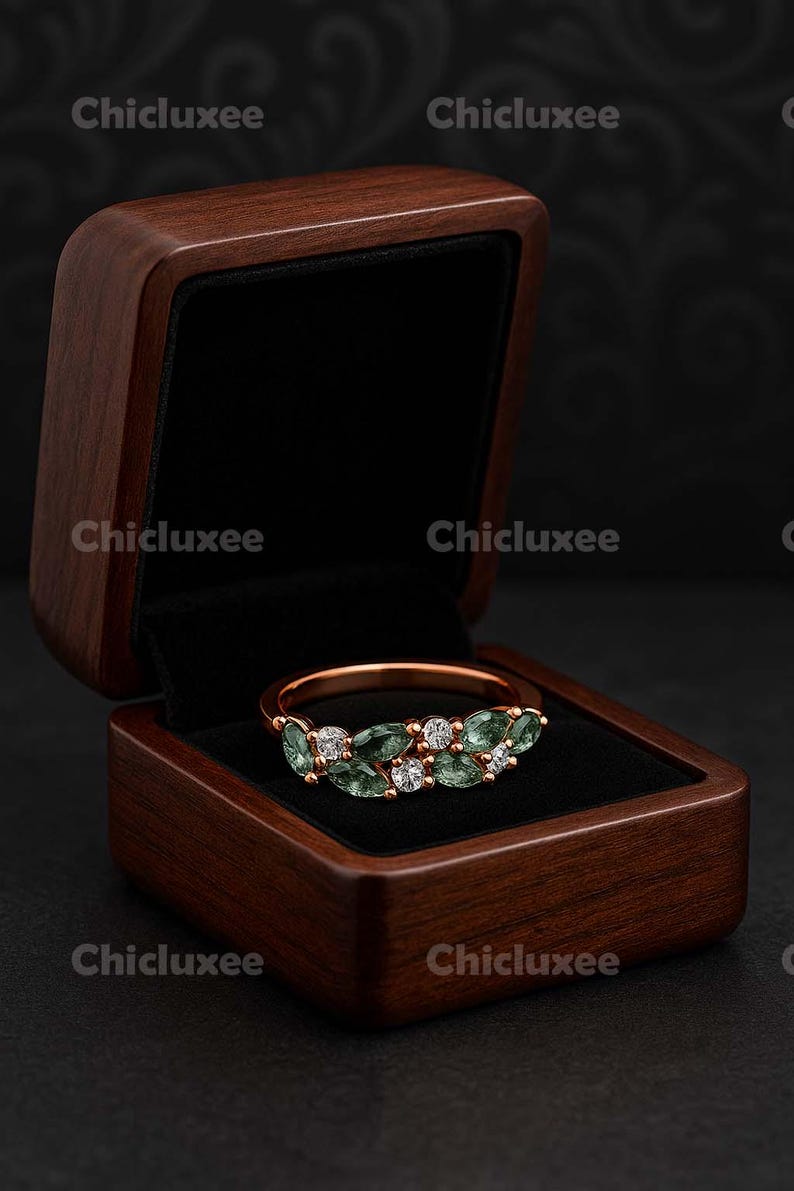 May include: A rose gold ring with green and white gemstones, displayed in an open wooden jewelry box with a black velvet interior. The box is a dark brown color. The brand name "Chicluxee" is visible.