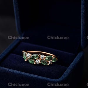 May include: A gold-toned ring with a leaf design, featuring green and clear gemstones. The ring is displayed in a dark blue velvet ring box. The gemstones are emerald green and clear, set in a delicate pattern.