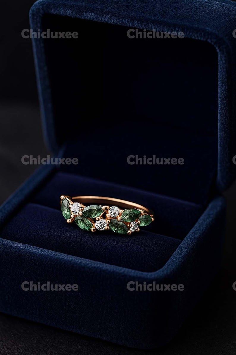May include: A gold-toned ring with a leaf design, featuring green and clear gemstones. The ring is displayed in a dark blue velvet ring box. The gemstones are emerald green and clear, set in a delicate pattern.
