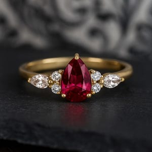 May include: A gold ring featuring a large, pear-shaped, deep red gemstone as the centerpiece. The ring is accented with smaller, round, and marquise-cut white gemstones. The band is a simple, polished gold.
