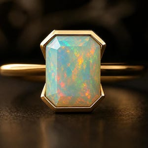 May include: A gold ring featuring a large, rectangular opal gemstone. The opal displays a play of color with hues of blue, green, and orange. The ring's band and setting are a polished gold color, creating a striking contrast.