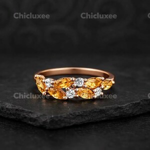 Puede incluir: Un anillo de oro rosa con un diseño de piedras preciosas amarillas y blancas. El anillo tiene una banda de oro rosa con una fila de piedras preciosas amarillas y piedras preciosas blancas. El anillo está sobre un fondo oscuro.
