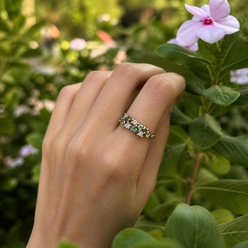 May include: A gold ring featuring a cluster of green and white gemstones. The ring is held up by a hand, with a blurred background of green foliage and a pink flower.