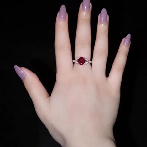 May include: A silver-toned ring featuring a round, deep red gemstone at the center, flanked by small, leaf-shaped clear stones on each side of the band. The ring is worn on a finger with light skin and long, almond-shaped mauve-colored fingernails.