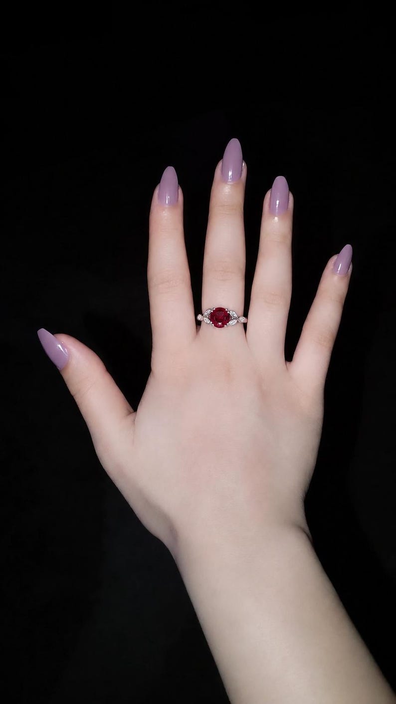 May include: A silver-toned ring featuring a round, deep red gemstone at the center, flanked by small, leaf-shaped clear stones on each side of the band. The ring is worn on a finger with light skin and long, almond-shaped mauve-colored fingernails.