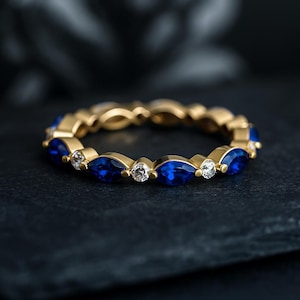 Op de afbeelding: Een gouden ring met een herhalend patroon van blauwe marquise-geslepen edelstenen en kleine witte edelstenen. De ring is geplaatst tegen een donkere achtergrond, waardoor de glans van de edelstenen wordt benadrukt.