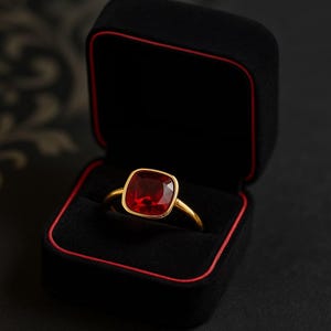 K&ouml;nnte beinhalten: Ein goldener Ring mit einem gro&szlig;en, quadratischen, roten Edelstein wird in einer offenen schwarzen Schmuckschatulle mit roter Umrandung pr&auml;sentiert. Der Ring hat ein d&uuml;nnes, goldenes Band, das das leuchtende Rot des Edelsteins erg&auml;nzt.