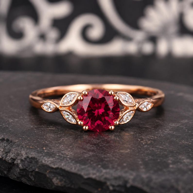 May include: A rose gold ring featuring a round, deep red center stone flanked by two marquise-cut clear stones on each side. The band is polished rose gold, and the setting holds the stones in a leaf-like arrangement along the ring's shank.