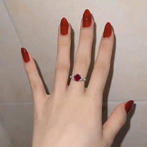 May include: A silver ring featuring a round, deep red gemstone at the center, flanked by small, clear, sparkling stones on each side of the band. The ring is worn on a finger, showcasing the contrast between the metallic band and the red stone.