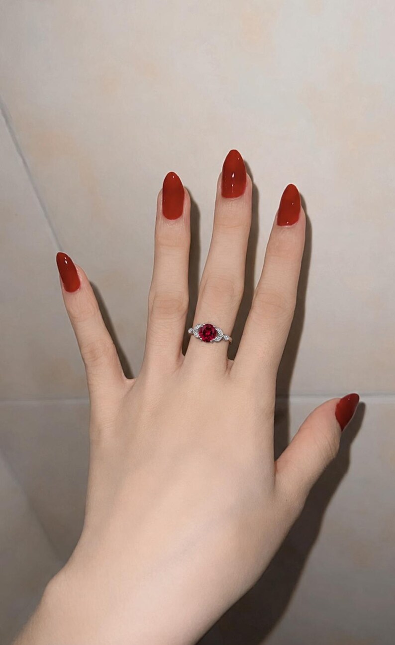 May include: A silver ring featuring a round, deep red gemstone at the center, flanked by small, clear, sparkling stones on each side of the band. The ring is worn on a finger, showcasing the contrast between the metallic band and the red stone.