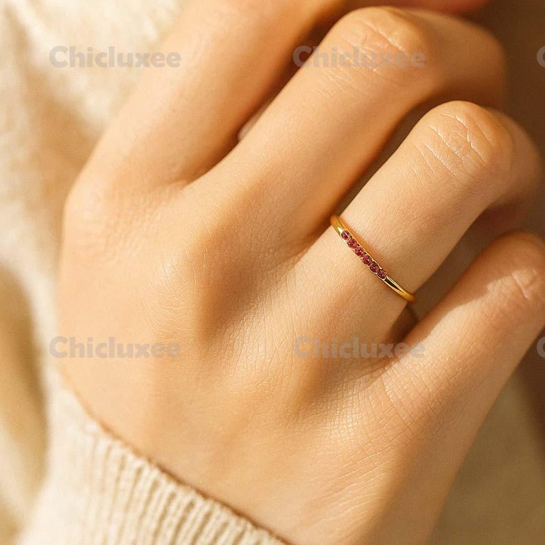May include: A gold ring with a row of small, red gemstones set across the top. The ring is on a person's finger, with the hand and part of the arm visible. The ring is a simple, elegant design.