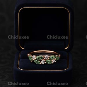 May include: A rose gold ring with green and clear gemstones, displayed in a navy blue velvet ring box. The ring features a series of oval-shaped green stones and small clear stones. The ring box is open, revealing the ring against a dark background.