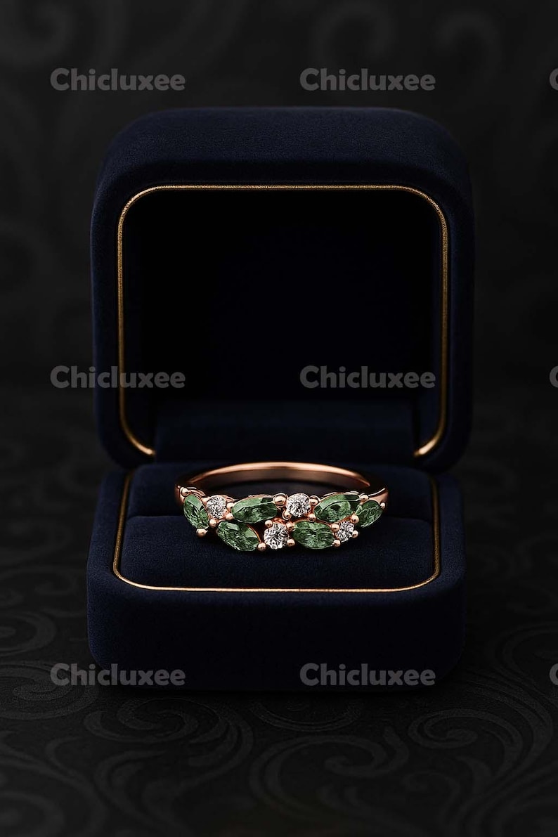 May include: A rose gold ring with green and clear gemstones, displayed in a navy blue velvet ring box. The ring features a series of oval-shaped green stones and small clear stones. The ring box is open, revealing the ring against a dark background.