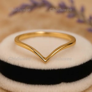 May include: A gold-colored V-shaped ring. The ring is set on a white and black surface. The ring's design is simple and elegant, with a smooth, polished finish. The ring is likely made of gold or a gold-colored metal.
