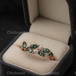May include: Rose gold ring with alternating green and clear gemstones. The ring is set in a velvet jewelry box. The gemstones are oval-shaped and arranged in a leaf-like pattern.