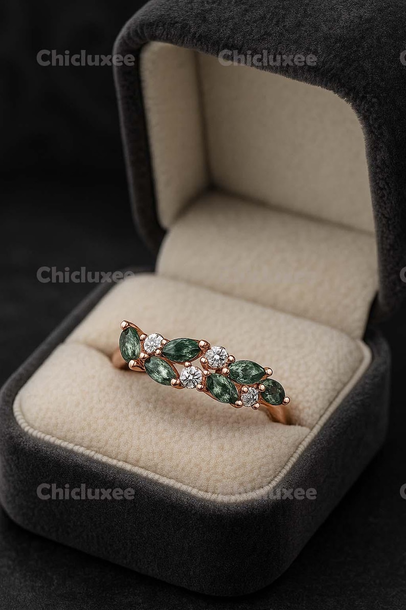 May include: Rose gold ring with alternating green and clear gemstones. The ring is set in a velvet jewelry box. The gemstones are oval-shaped and arranged in a leaf-like pattern.