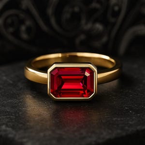 May include: A gold ring featuring a rectangular, deep red gemstone. The gemstone is set in a bezel setting, and the band is a simple, polished gold. The ring is displayed on a dark, textured surface.