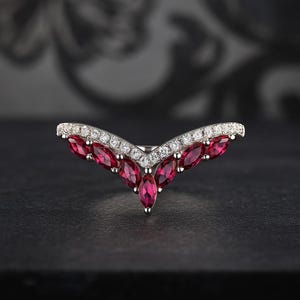 May include: A silver ring with a V-shaped design. The ring features a row of small, clear stones along the top and a series of marquise-cut, deep red gemstones in the center. The ring is set against a dark background.