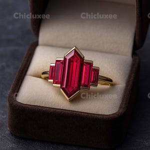 May include: A gold ring with a large, rectangular, red gemstone at the center, flanked by smaller rectangular red stones. The ring is set in a brown velvet box. The brand name "Chicluxee" is visible.